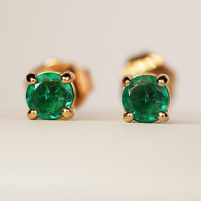 Close up of a pair of emerald earrings in a gold four prong setting, set against a neutral background.
