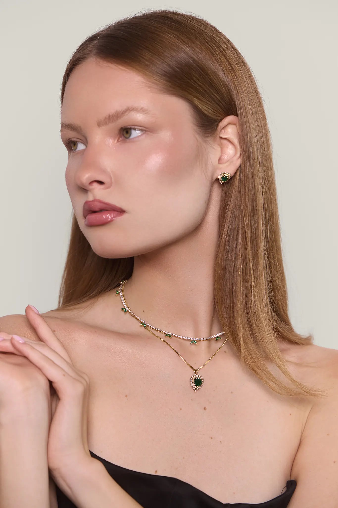 Woman in a black top wearing diamond and emerald heart shaped earrings and a matching necklace along with a diamond tennis necklace