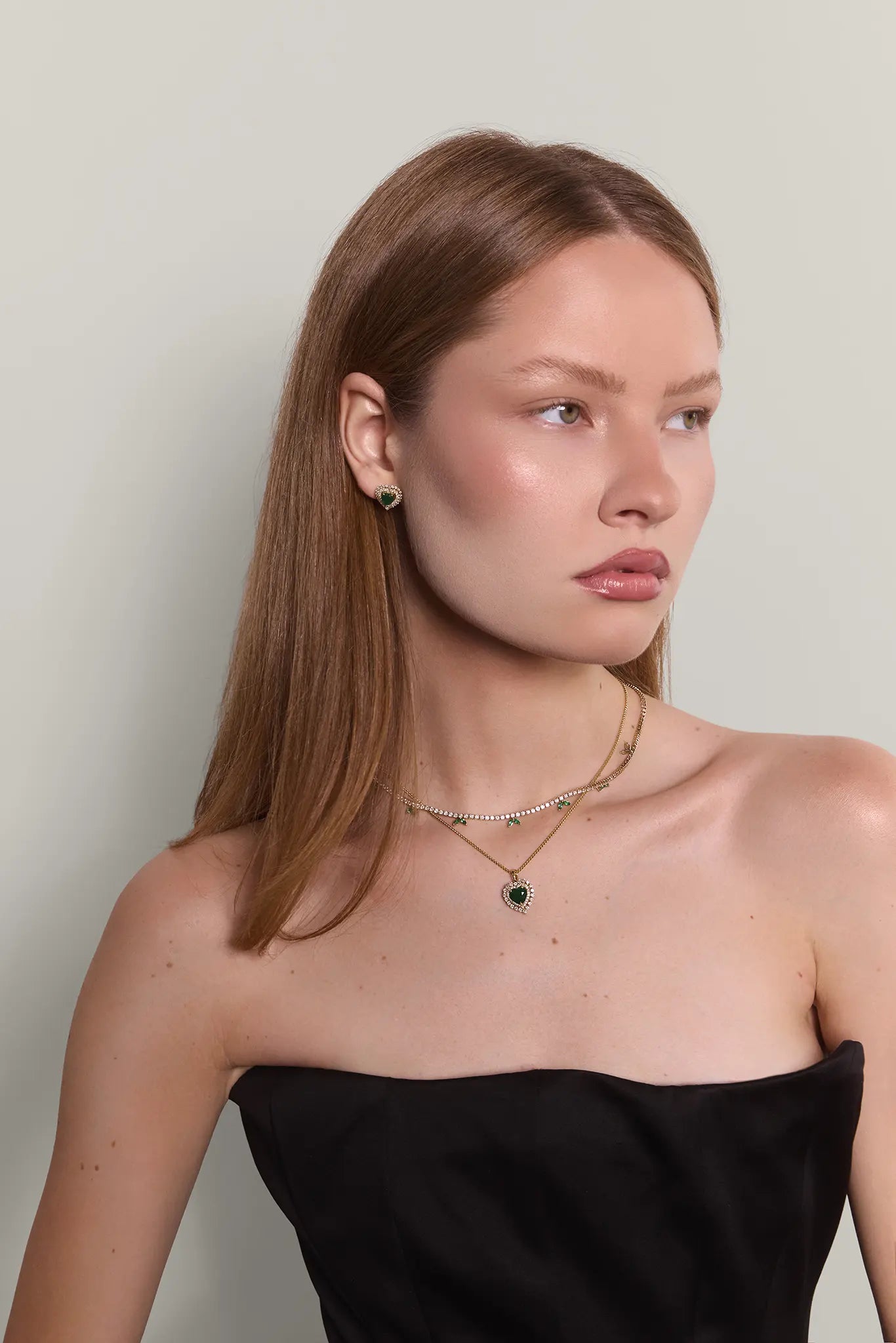 Woman wearing a black strapless top and jewelry comprising heart shaped emerald earrings, a matching necklace and a diamond tennis necklace
