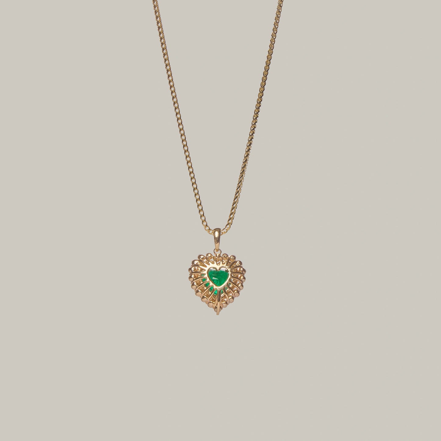 Rear-facing close-up of a gold necklace with a heart-shaped emerald pendant on a light gray background