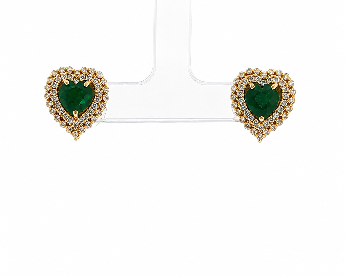 A pair of heart-shaped stud earrings with emerald-cut green centers, surrounded by gold and clear stones, displayed on a clear stand against a white background.