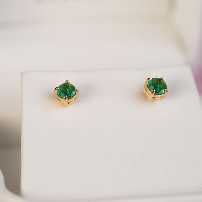 Top-down shot of a pair of emerald earrings in a gold four prong setting, set against a neutral background.