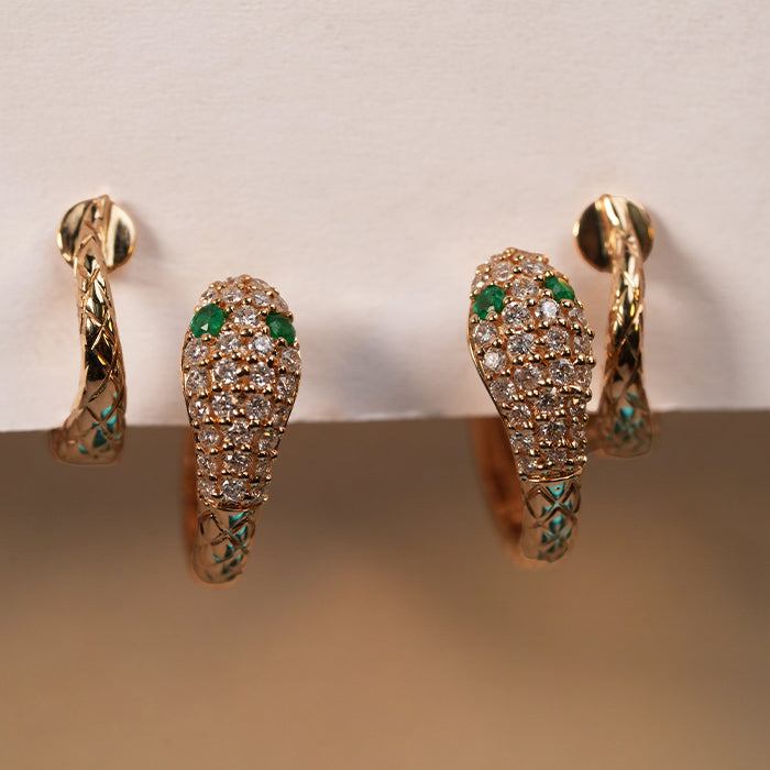 A pair of gold snake earrings with emerald eyes and pave set diamonds against a neutral background.