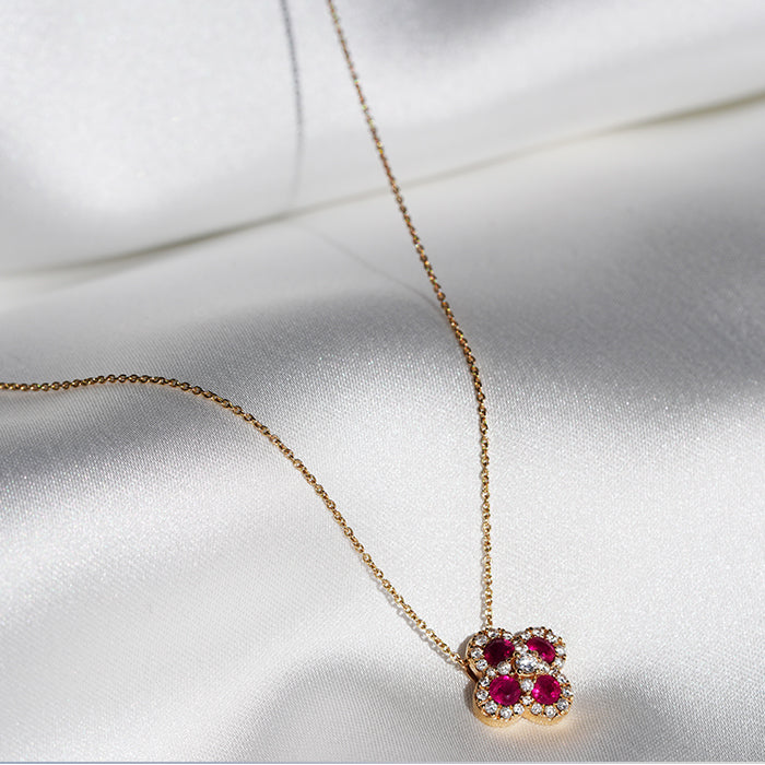 A gold clover shaped pendant with four rubies, a central diamond and pave set diamonds around the outside, hanging from a gold chain and draped against a white fabric background.