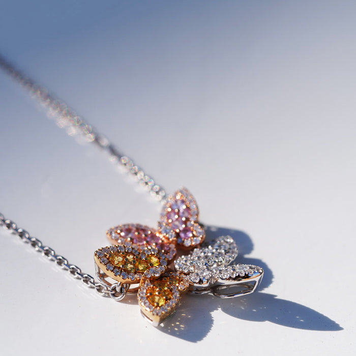 A diamond-studded necklace featuring three butterfly-shaped pendants in gold, pink and classic white with pave-set stones against a neutral background.