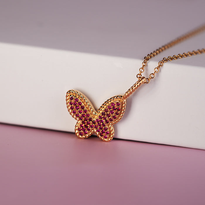A gold butterfly pendant adorned with rubies, displayed on a white box with a pink backdrop.