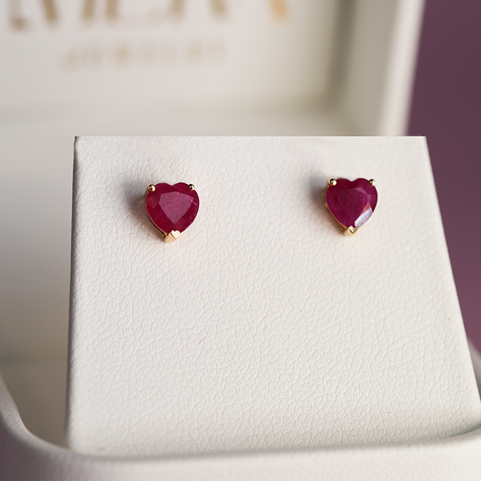 A pair of heart-shaped ruby earrings set in 14k gold, displayed on a white stand.
