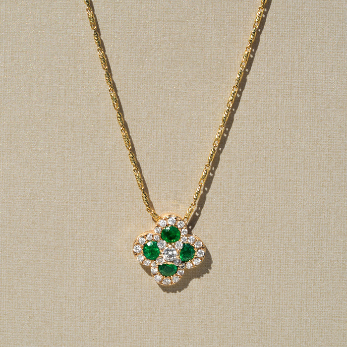 A gold clover shaped pendant with four emeralds, a central diamond and pave set diamonds around the outside displayed on a neutral textured background.