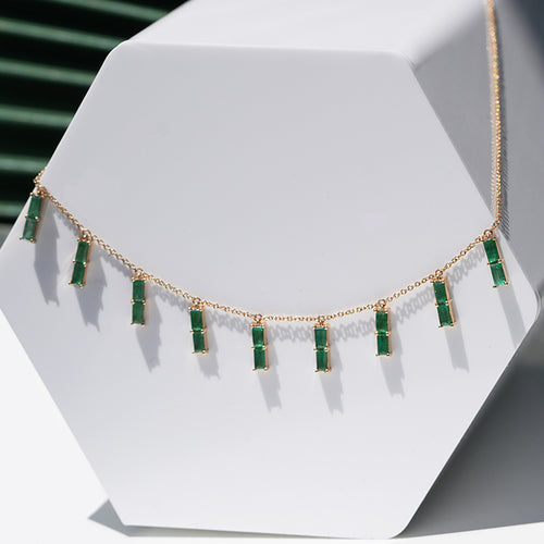 A gold necklace with vertical cut dangling emeralds showcased on a white box against a textured green background.