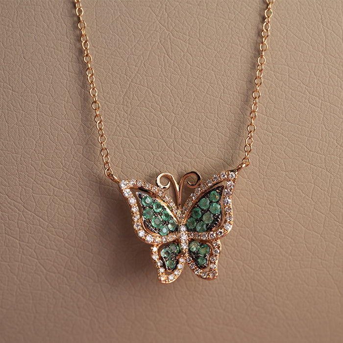 Close up of a gold butterfly pendant with emerald wings and encrusted diamonds suspended on a chain against a textured cream backdrop.