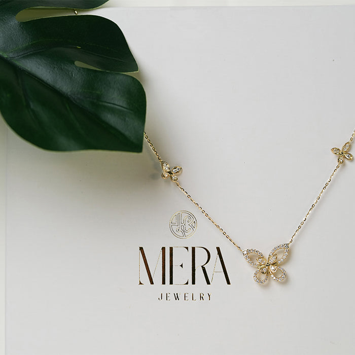 A gold-colored necklace featuring three butterfly-shaped charms with intricate detailing, displayed on a white background with a green leaf.