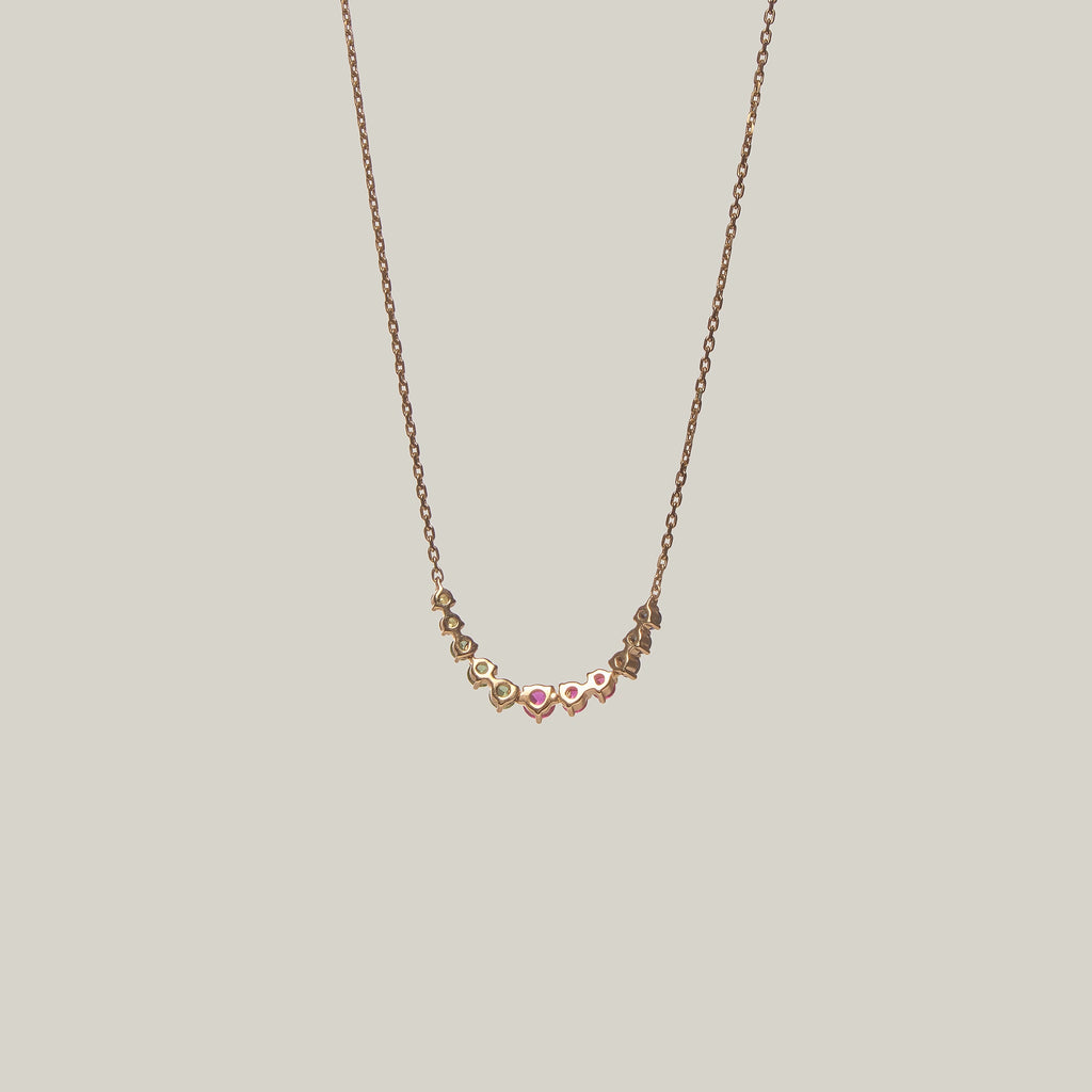 Close-up rear-facing view of a delicate curved necklace with multicolored gemstones in violet, green, yellow and pink on a light background.