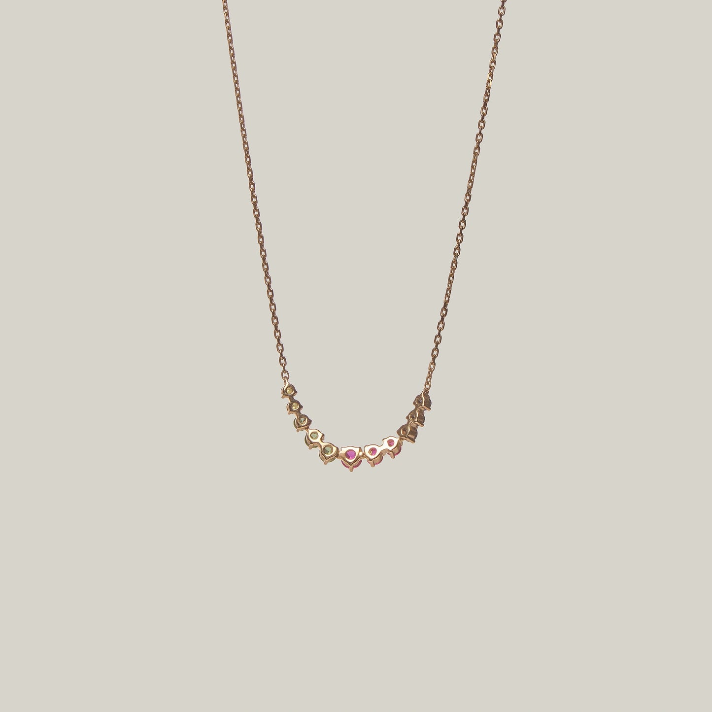 Close-up rear-facing view of a delicate curved necklace with multicolored gemstones in violet, green, yellow and pink on a light background.