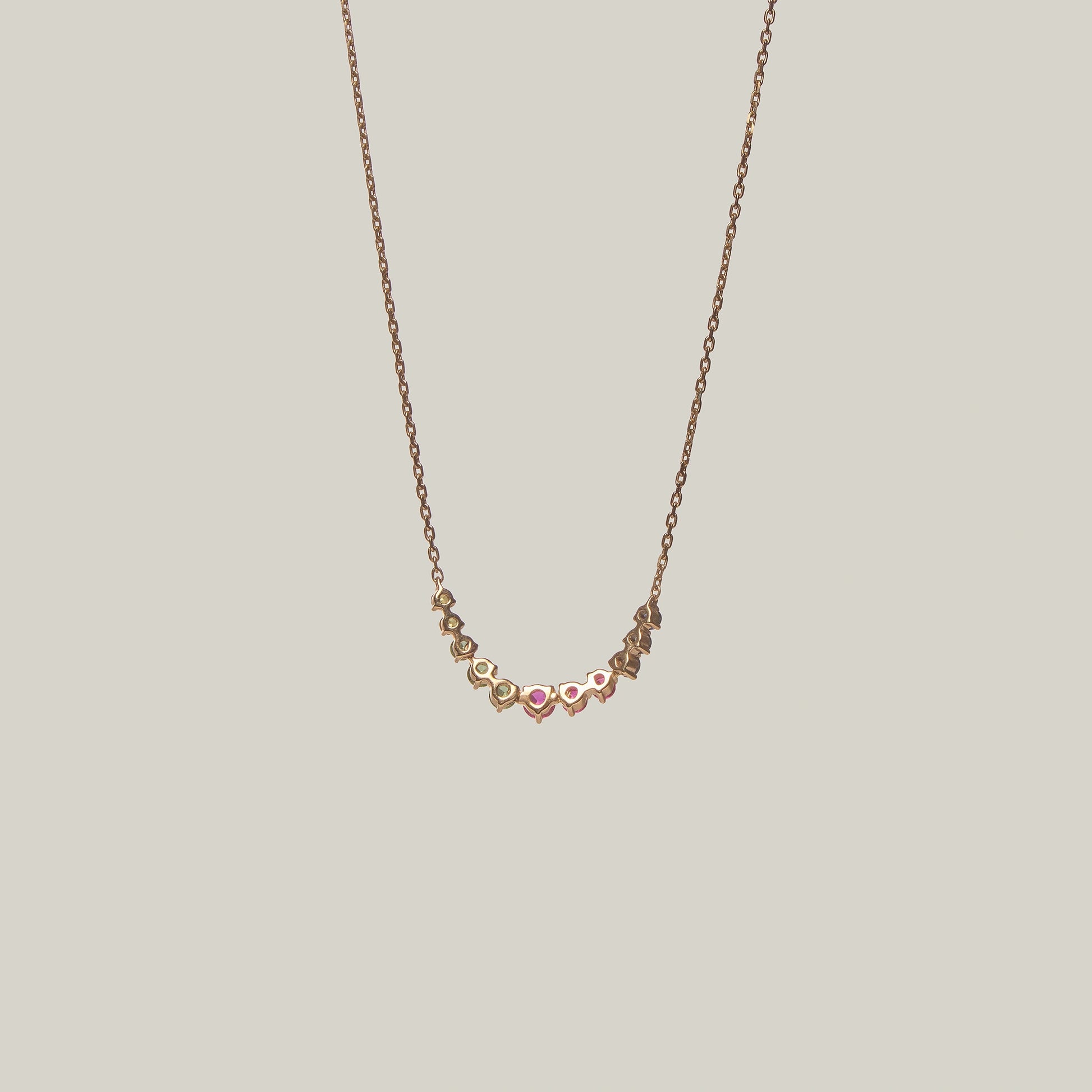 Close-up rear-facing view of a delicate curved necklace with multicolored gemstones in violet, green, yellow and pink on a light background.