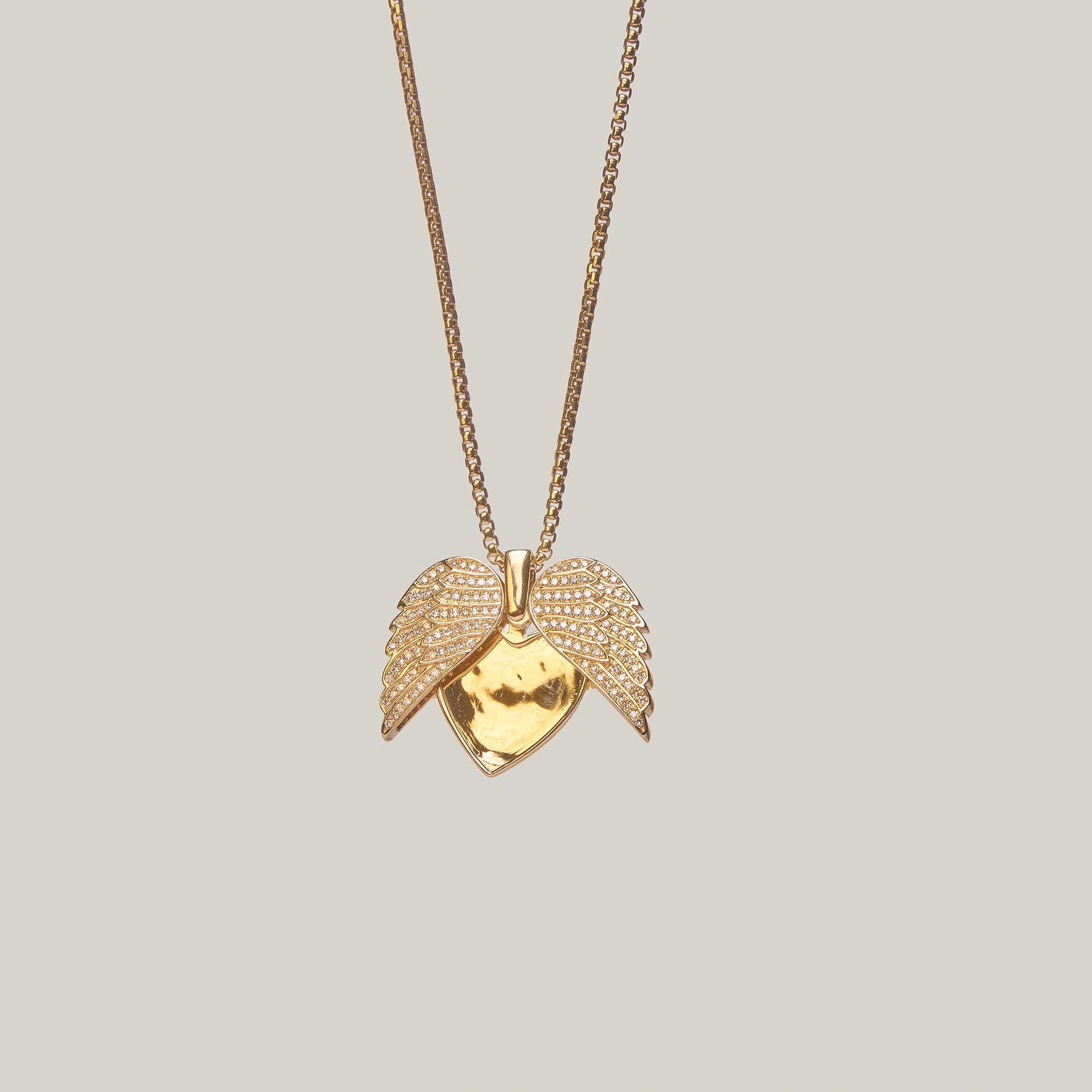 Close-up front-facing view of a gold heart pendant with an opened angel wings design, encrusted with diamonds, on a light gray background