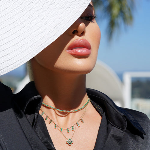 A woman wearing a white hat and a black shirt, wearing 3 emerald necklaces. One is a tennis necklace, one features vertical cut emeralds and one is in a clover shape.
