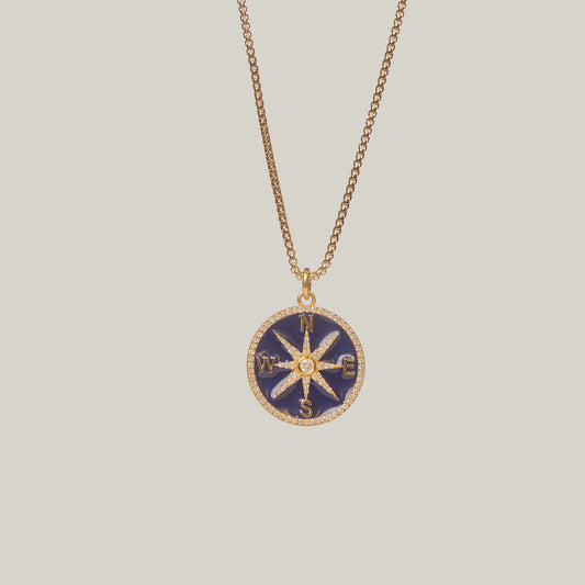 Close up front facing view of a gold necklace with a compass pendant on a light gray background