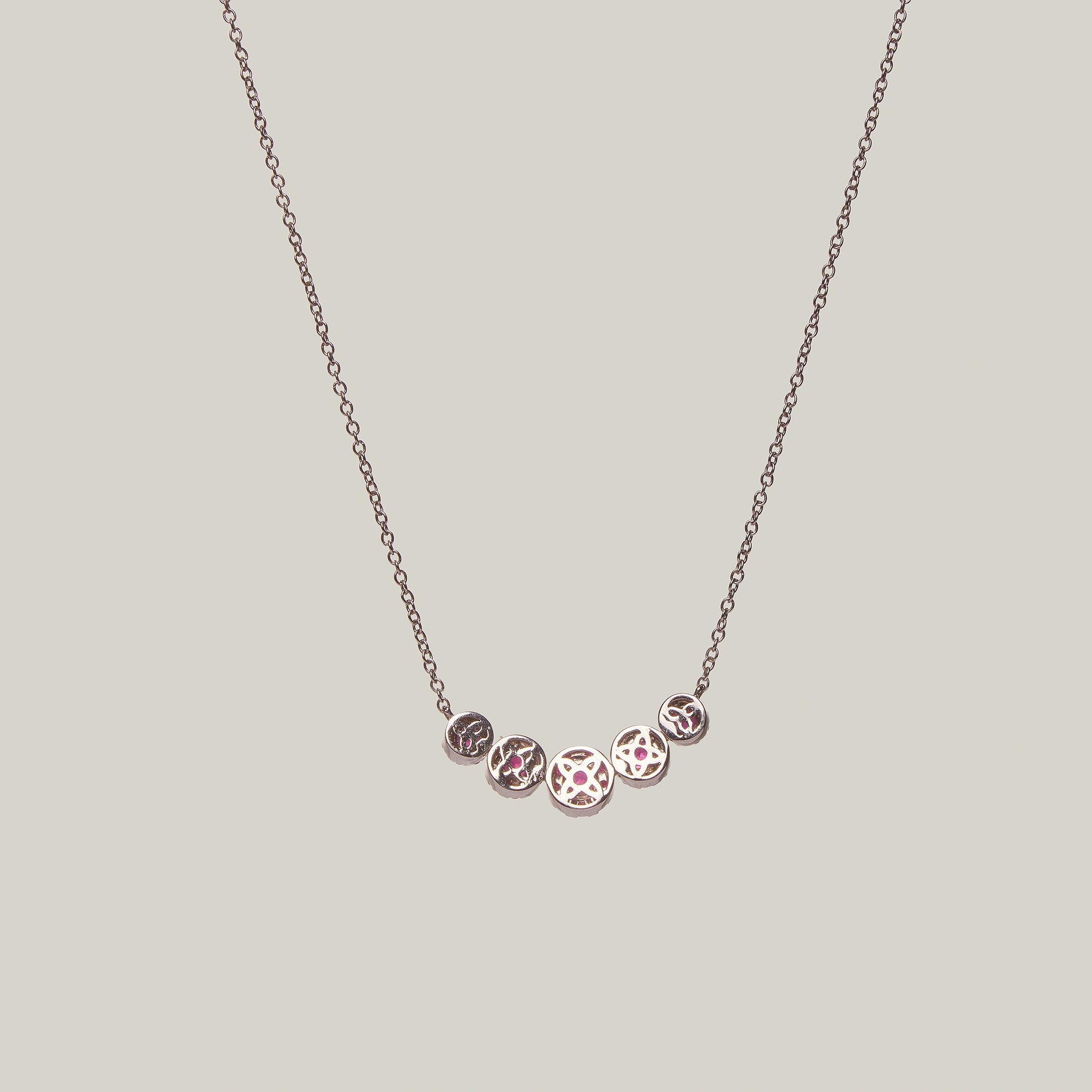 Rear view of a gold necklace with rubies in a curved pattern on a light gray background