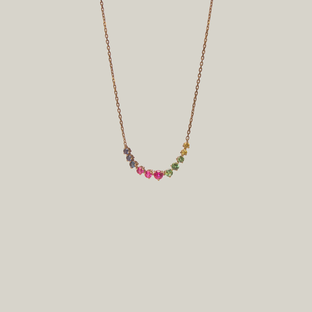 Close-up front-facing view of a delicate curved necklace with multicolored gemstones in violet, green, yellow and pink on a light background.