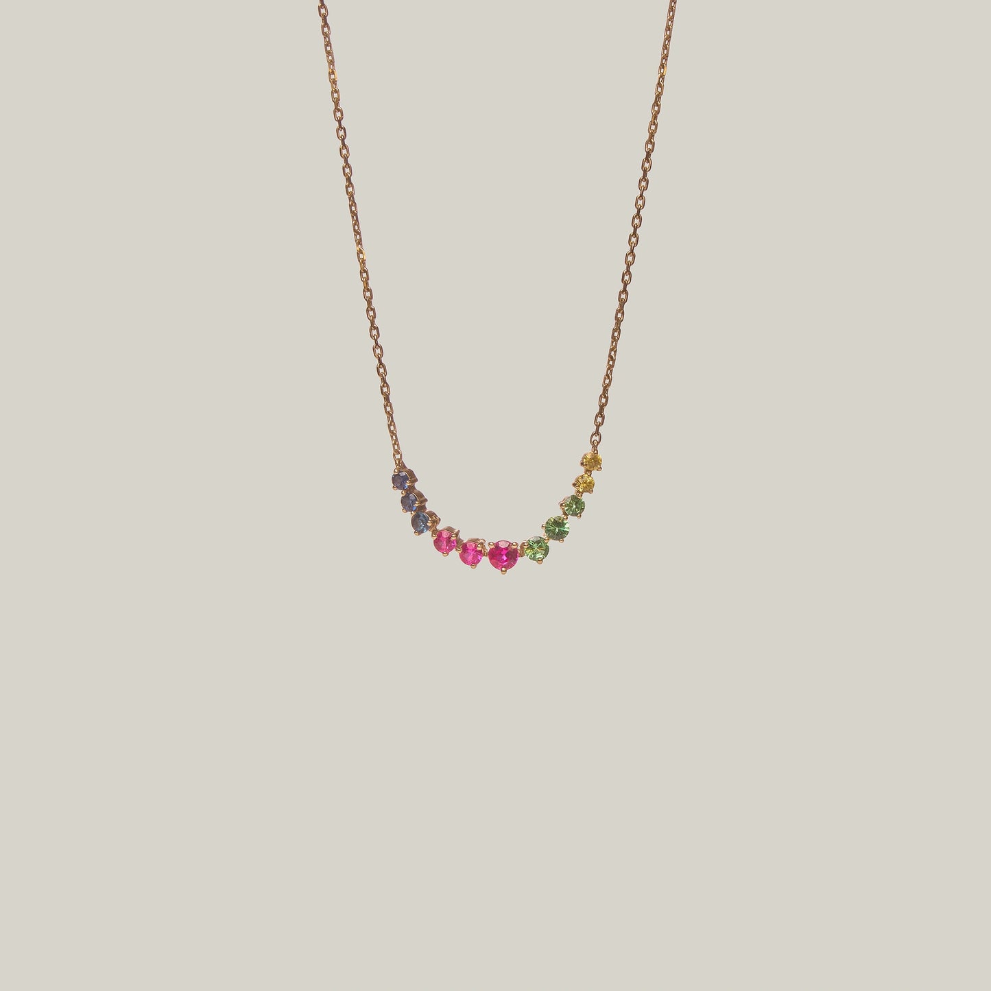 Close-up front-facing view of a delicate curved necklace with multicolored gemstones in violet, green, yellow and pink on a light background.