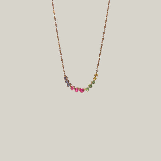 Close-up front-facing view of a delicate curved necklace with multicolored gemstones in violet, green, yellow and pink on a light background.