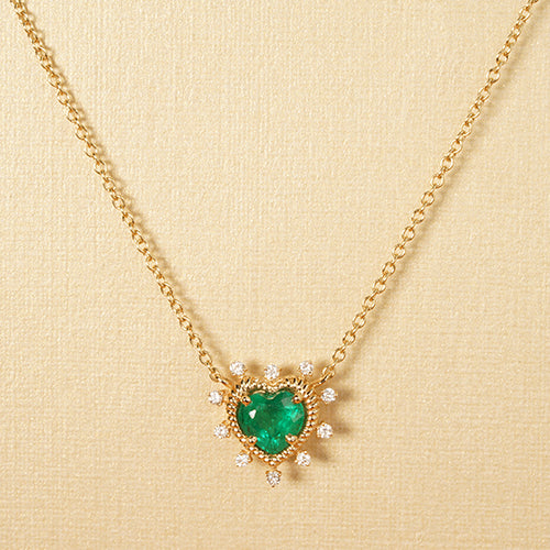 A gold necklace featuring a central emerald and diamond accents, suspended from a chain against a textured white background.