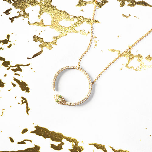A gold-colored pendant necklace with a snake design, featuring white diamonds and emeralds, displayed on a white and gold textured background.