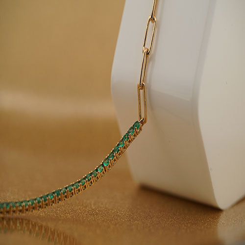 A close-up image of a gold bracelet with emeralds, displaying the detailed chain design and the sparkling stones.