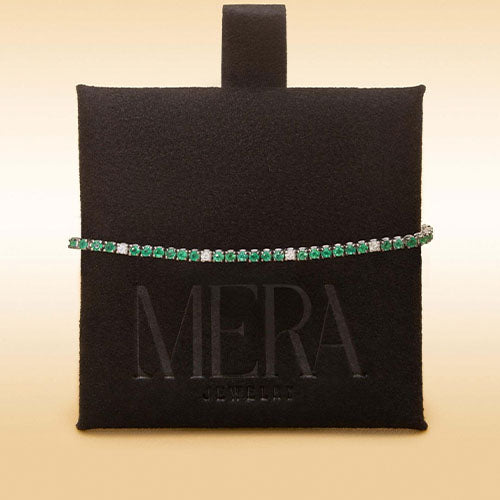 A diamond and emerald tennis bracelet displayed on a black cushion against a cream background.