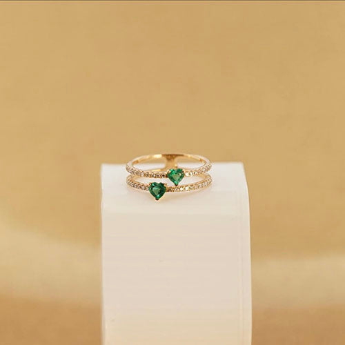 A gold ring with an emerald centerpiece, featuring encrusted diamonds and positioned on a white block in front of a dark cream background.