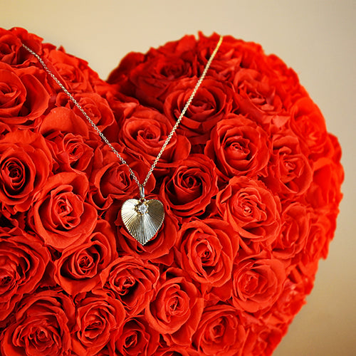 A 14k gold heart-shaped pendant with a diamond centerpiece, displayed on a bed of red roses.