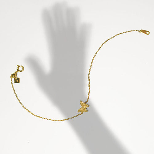 A gold bracelet with a single butterfly charm and hand set diamonds against a white backdrop with the silhouette of a hand to illustrate its length.