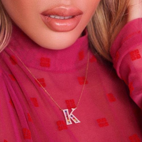 Model wears a pink top with red squares and a necklace in the letter K with encrusted diamonds outlining the shape and multicolored sapphires in the center.