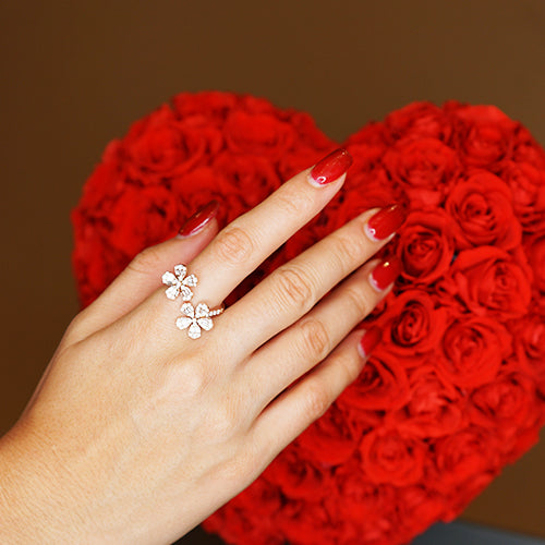 Hand wearing red nail polish and a gold diamond ring with two flowers against a backdrop of a large heart comprised of red roses.