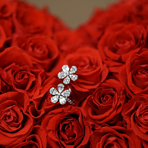 A close-up view of a gold ring with two diamond flowers, displayed against a backdrop of red roses.