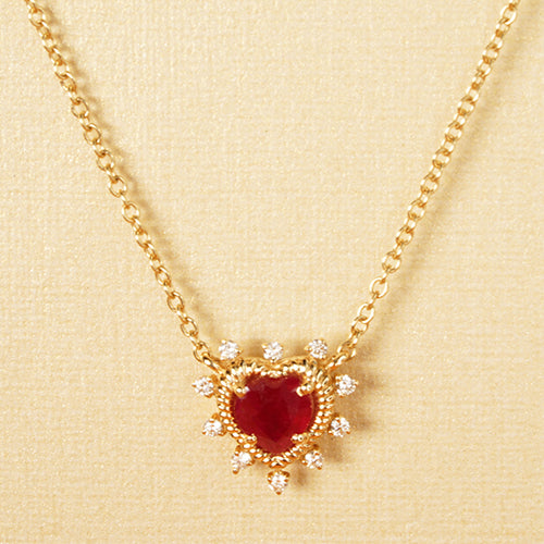 A gold necklace with a central ruby heart and diamond accents, suspended from a gold chain against a cream textured background.
