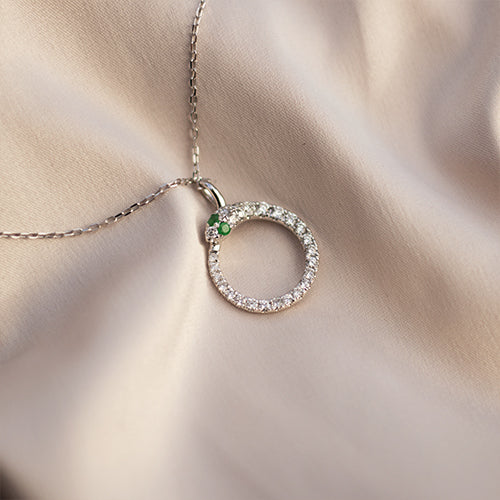 A silver-colored pendant necklace with a snake design, featuring white diamonds and emeralds, displayed on a soft fabric surface.